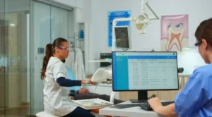 Dental clinic staff reviewing patient records on a computer while a dentist prepares equipment in the operatory
