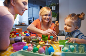 Two women and a little girl painting colorful Easter eggs at a kitchen table with paint and brushes