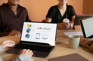 People in a meeting reviewing an analytics dashboard on a laptop with charts, tablets, and coffee cups