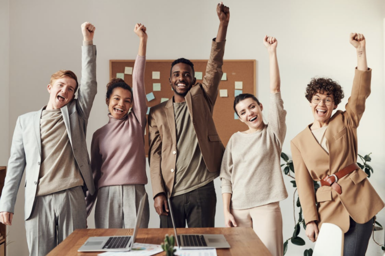 Happy diverse team in casual office attire raises arms in celebration around a table with laptops and notes
