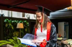 Young woman in graduation cap and gown reading a folder indoors, smiling in a modern lounge area