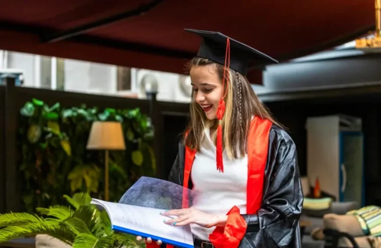 Young woman in graduation cap and gown reading a folder indoors, smiling in a modern lounge area