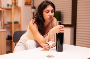 Upset woman sitting at a table holding a wine bottle with an empty glass in a home setting