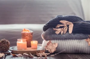 Cozy home setup with lit candles, folded knit blankets, and autumn leaves on a wooden table