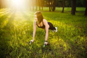 Woman exercising outdoors doing push-ups with handles on green grass in a sunny park