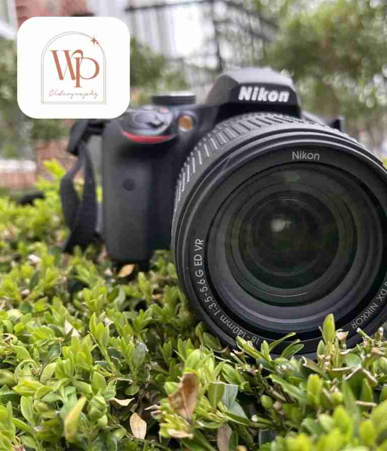Close-up of a Nikon DSLR on green shrubs, wide lens facing camera, with WP Photography logo overlay