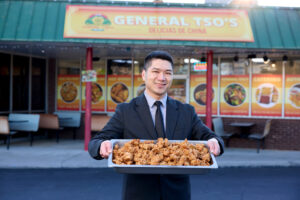 Smiling man in a suit holds a tray of fried chicken outside a restaurant storefront with signage in the background