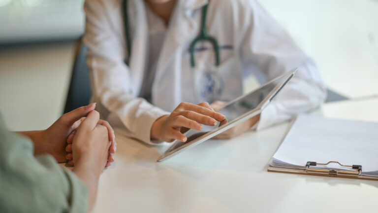 Doctor in a white coat uses a tablet while speaking with a patient at a clinic desk, with a clipboard nearby