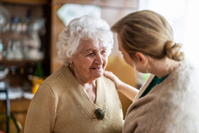 Caregiver talks with and supports an elderly woman at home, offering comfort and help with daily care