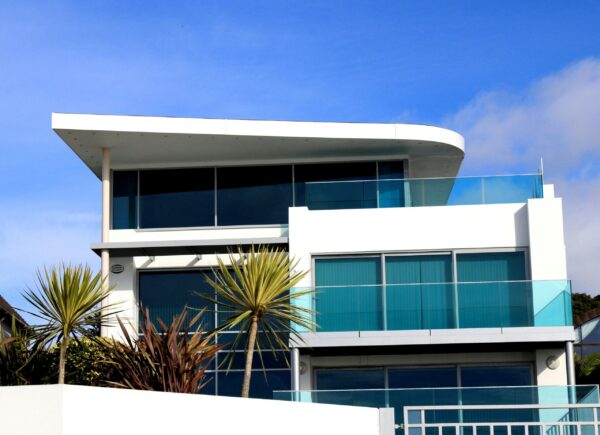 Modern white home with large windows and glass balcony rails, with palm-like plants in front and blue sky above