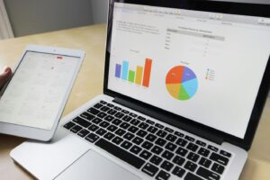 Laptop displays charts and a data table on screen, with a tablet showing a calendar on a desk beside it