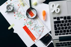 Desk with printed stock charts, a magnifying glass, calculator, laptop keyboard, and a percent symbol for finance work