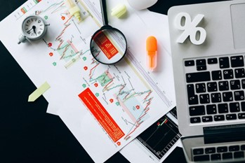 Desk with printed stock charts, a magnifying glass, calculator, laptop keyboard, and a percent symbol for finance work